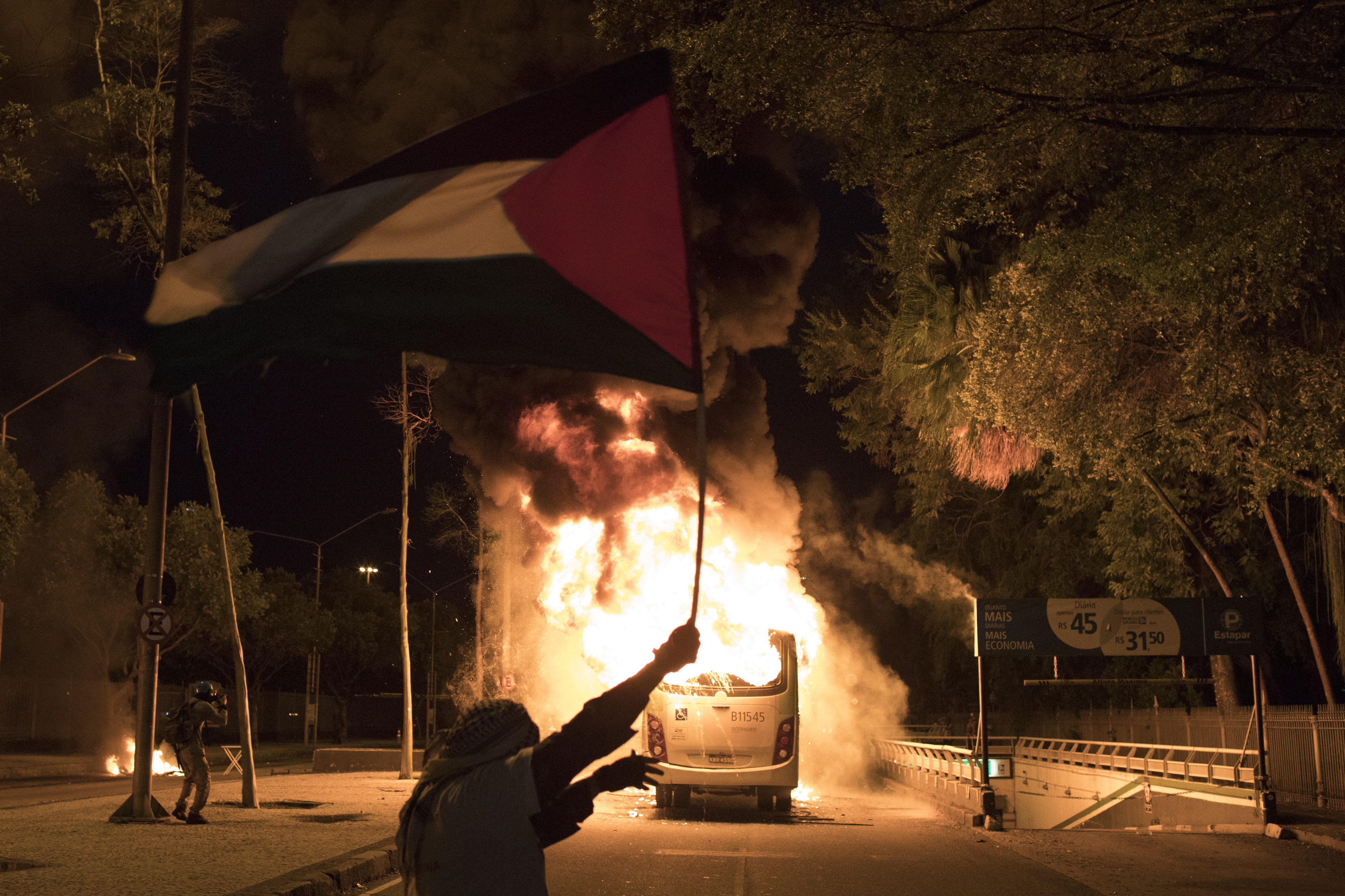 A masked demonstrator waves a Palestinian flag by a bus set fire by protesters after clashes with police broke out during a general strike in Rio de Janeiro, Brazil, Friday, April, 28, 2017. Public transport largely came to a halt across much of Brazil on Friday and protesters blocked roads and scuffled with police as part of a general strike to protest proposed changes to labor laws and the pension system. (AP Photo/Leo Correa)