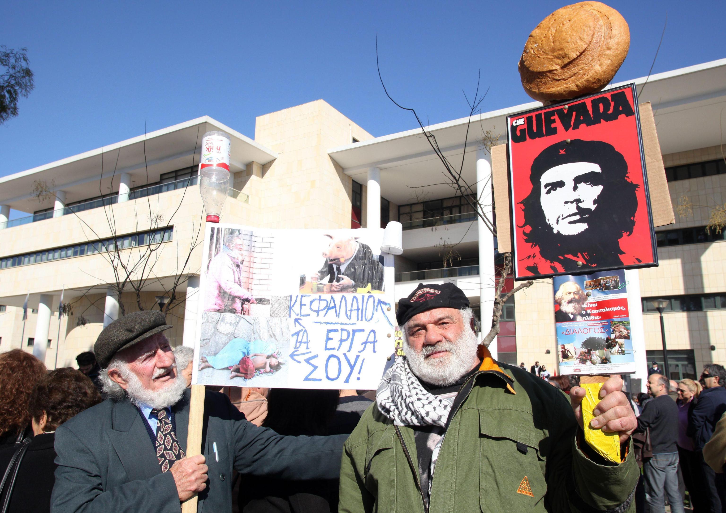 epa04061401 Protesters stand with placards, one reading 'Capital your deeds' (L) and the other showing a portrait of Argentinian Marxist revolutionary Che Guevara (R), during a demonstration outside the Ministry of Finance against the troika's austerity measures, in Nicosia, Cyprus, 08 February 2014. A protest against austerity measures imposed by the troika was held outside the Central Cooperation Bank as well as the Ministry of Finance.  EPA/KATIA CHRISTODOULOU