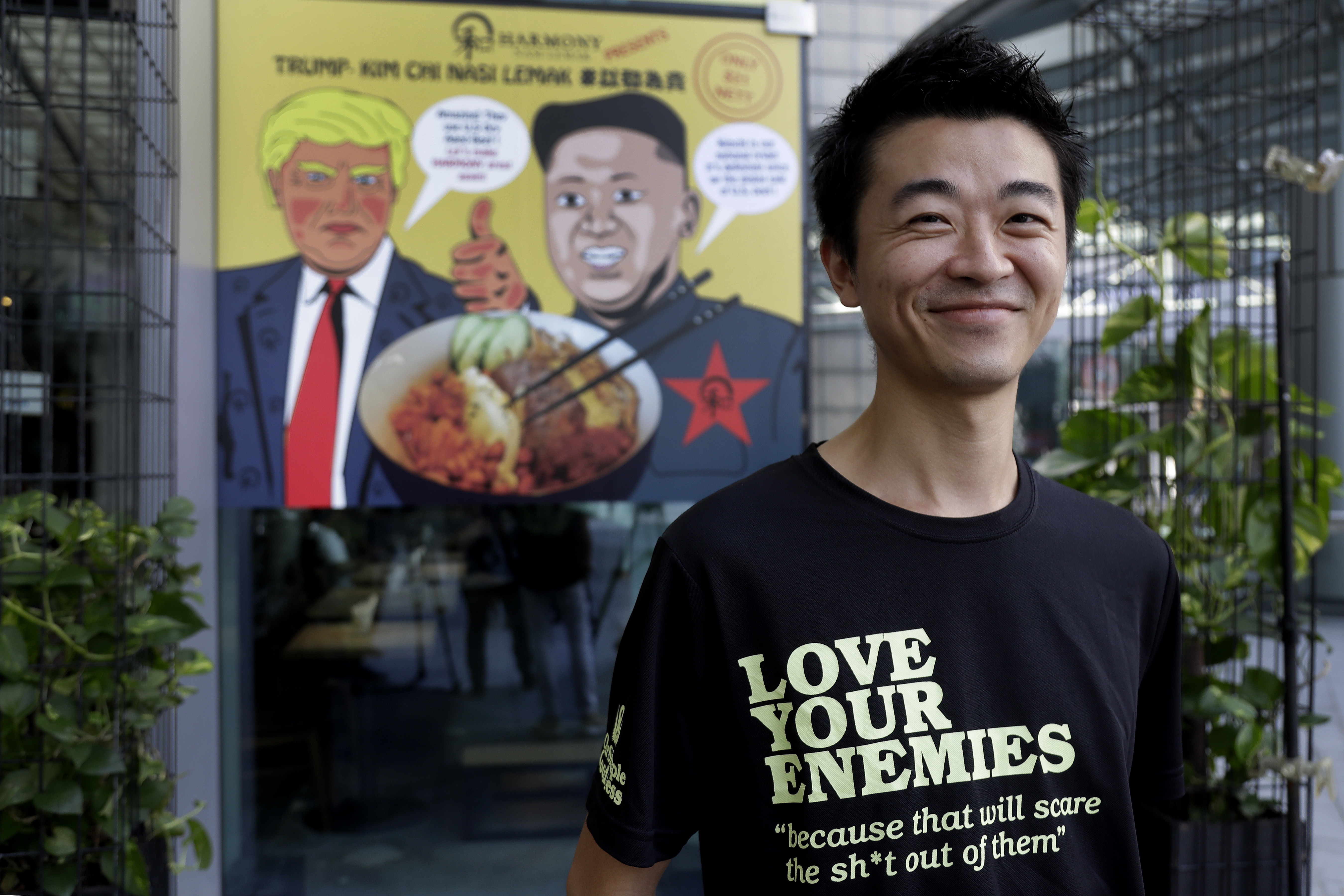 In this June 7, 2018, photo, Zach Wen, 34, co-founder of Harmony Nasi Lemak poses in front of an advertisement board of cartoon caricatures of U.S. President Donald Trump and North Korean leader Kim Jong Un which are supposed to be the inspiration behind a local dish, the 