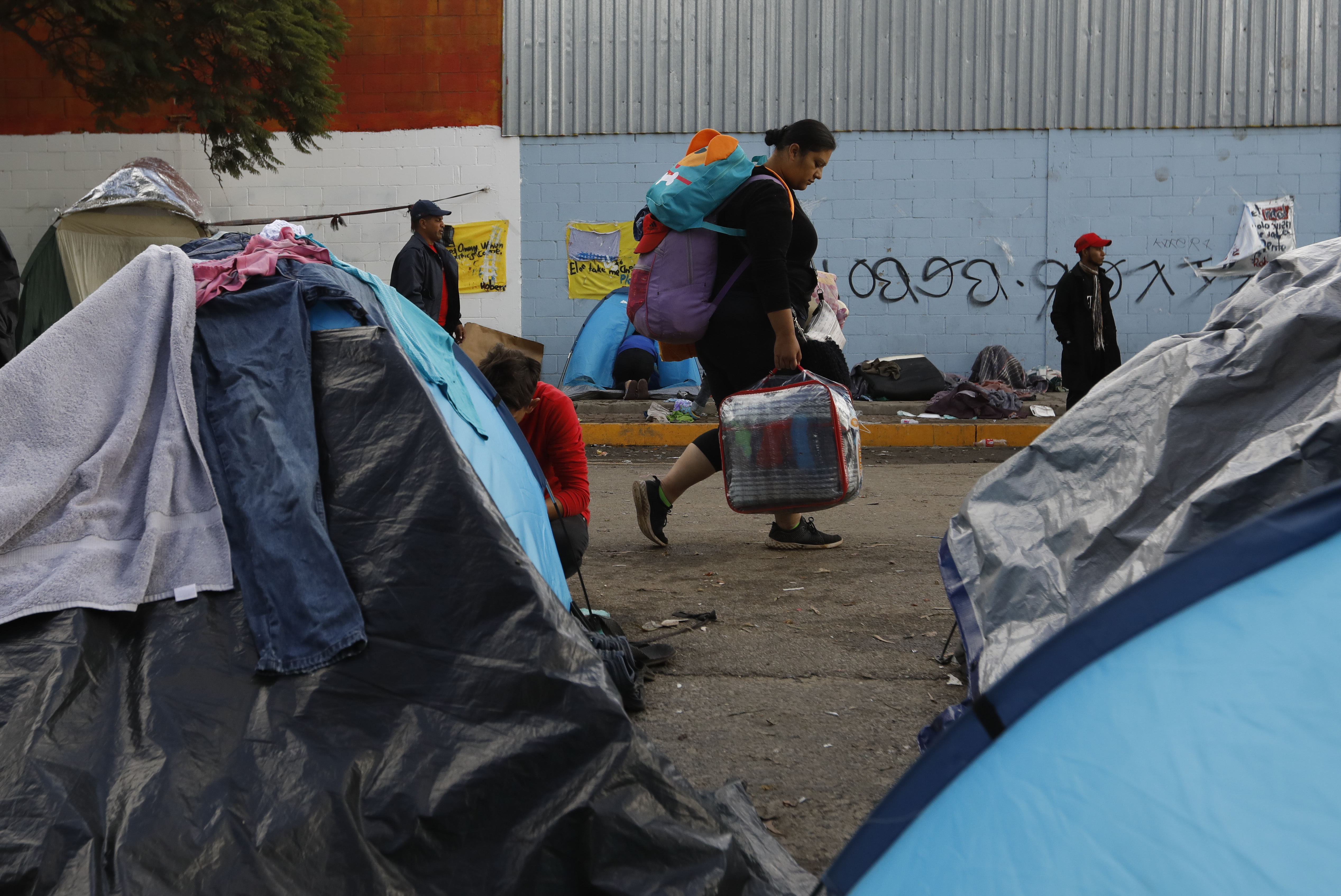 A woman carries her bags past tents as migrants decide individually whether to accept the city's plan to move them to a new shelter from the Benito Juarez sports complex, in Tijuana, Mexico, Friday, Nov. 30, 2018. Authorities in the Mexican city of Tijuana have begun moving some of more than 6,000 Central American migrants from an overcrowded shelter on the border to an events hall further away.(AP Photo/Rebecca Blackwell)