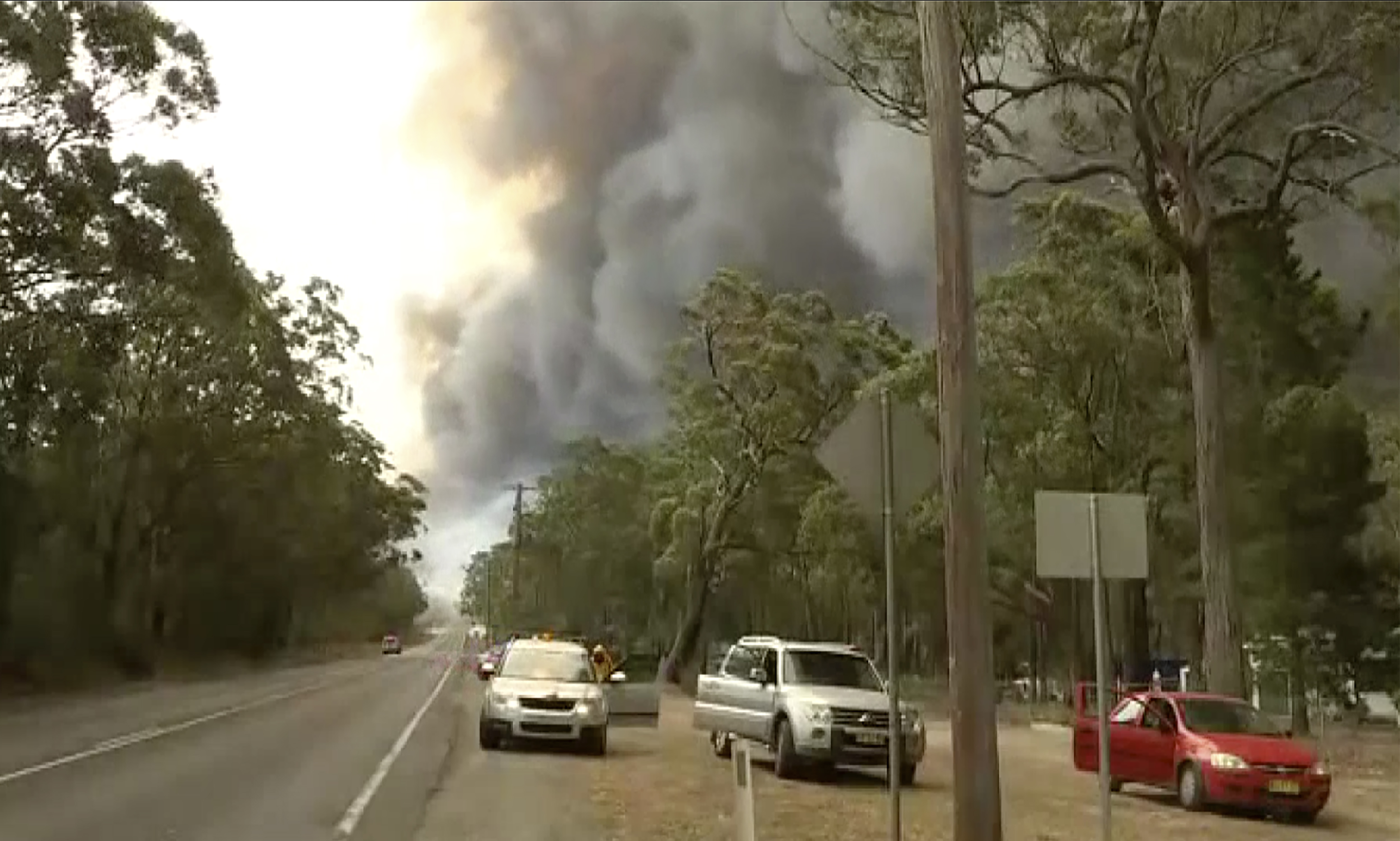 In this image made from video, vehicles are parked during a road closure as smoke rises in the rear, Tuesday, Dec. 31, 2019, in Brogo, new South Wales, Australia. Wildfires burning across Australia's two most-populous states have trapped residents of a seaside town in apocalyptic conditions, destroyed many properties and caused at least two fatalities. (Australian Broadcasting Corporation, Channel 9, Channel 7 via AP)