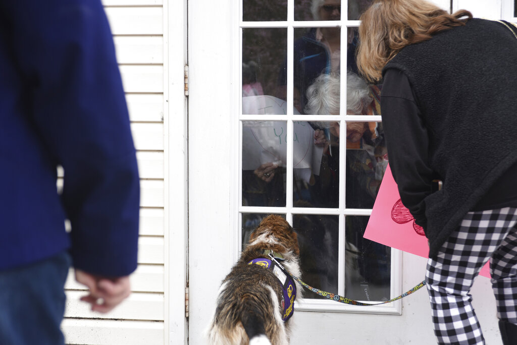 Under a cloudy sky on Thursday, March 26, 2020, a group of about 25 people, including members of the Alliance of Therapy Dogs and family members of residents, marched around Bright Leaf Senior Living, in Danville, Va., which has been closed to visitors for several weeks in an effort to curb the spread of COVID-19. Many of the social activities inside the facility have also been canceled, leaving the residents all but isolated in their rooms. The marchers smiled, waved, held up homemade signs and tried to talk to the residents inside, many of whom smiled and waved back and held up their own signs.  (Caleb Ayers/Danville Register & Bee via AP)