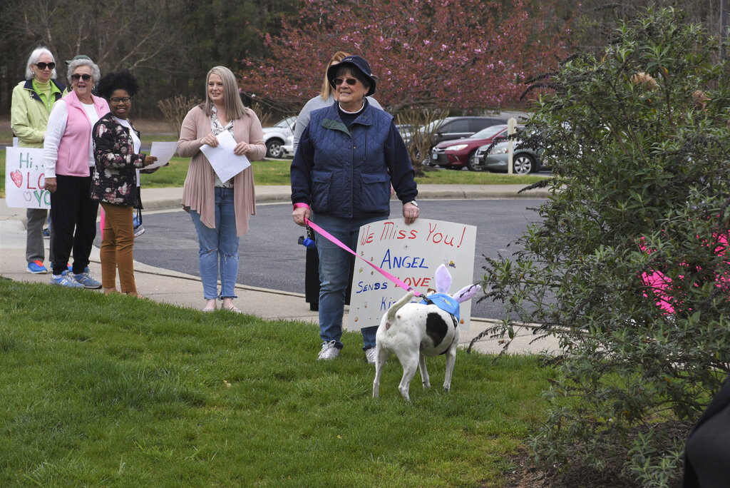 Under a cloudy sky on Thursday, March 26, 2020, a group of about 25 people, including members of the Alliance of Therapy Dogs and family members of residents, marched around Bright Leaf Senior Living, in Danville, Va., which has been closed to visitors for several weeks in an effort to curb the spread of COVID-19. Many of the social activities inside the facility have also been canceled, leaving the residents all but isolated in their rooms. The marchers smiled, waved, held up homemade signs and tried to talk to the residents inside, many of whom smiled and waved back and held up their own signs. (Caleb Ayers/Danville Register & Bee via AP)