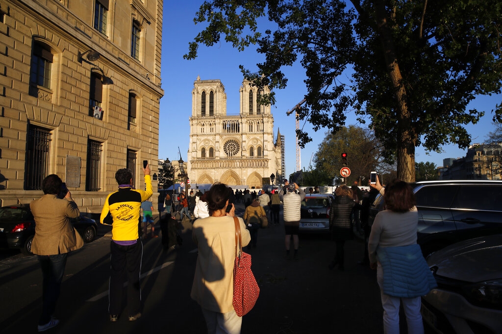 People stop in front of Notre Dame Cathedral as the bells ring to mark one year of blaze Wednesday, April 15, 2020 in Paris. A year after a devastating fire, the restoration of Notre Dame Cathedral has been halted by a lockdown in Paris to battle the coronavirus. The April 15, 2019, blaze gutted its interior, toppled its famous spire and horrified the world. (AP Photo/Christophe Ena)