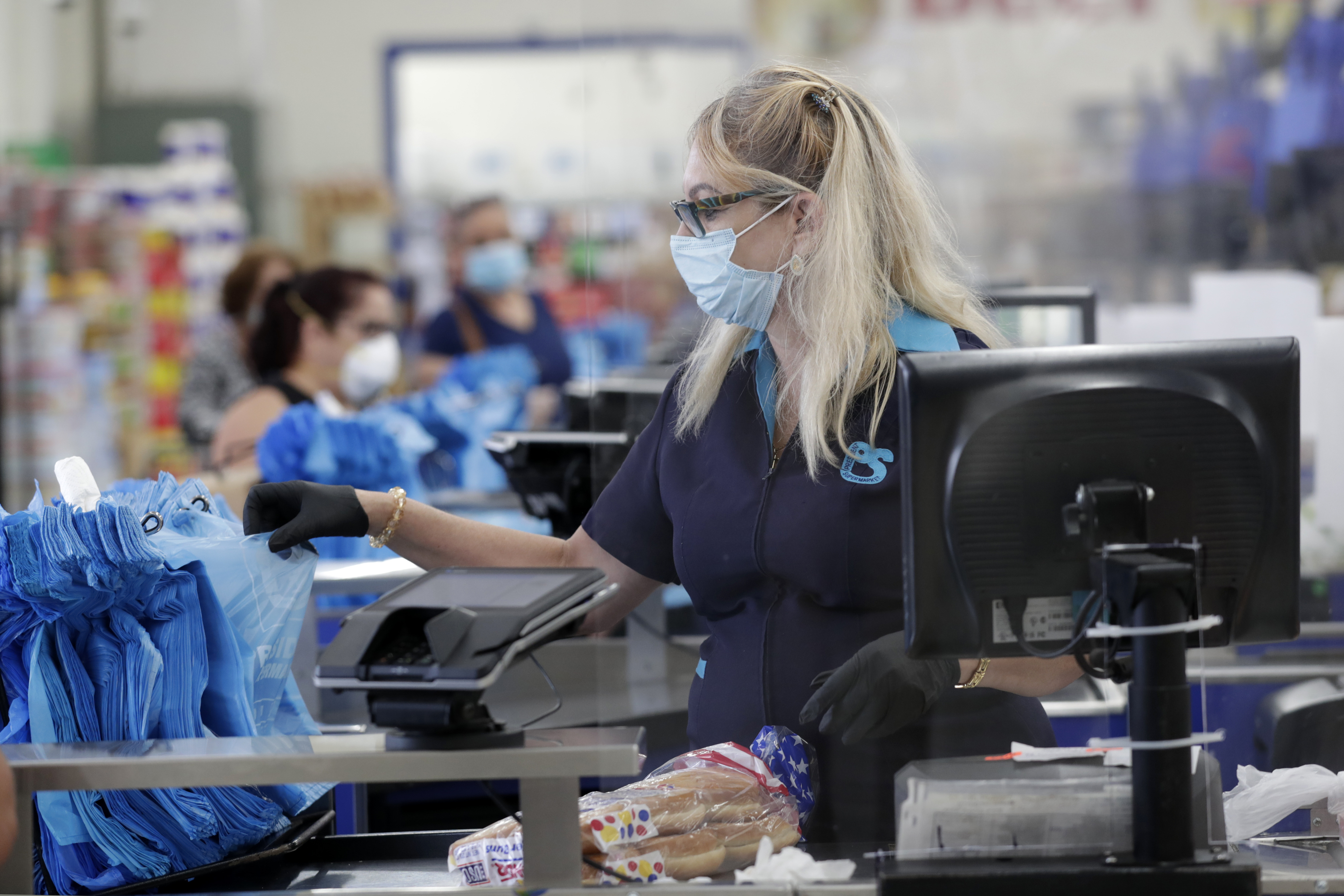 Cashier Mercedes Peña wears a protective mask as she works behind a plastic shield at the Presidente Supermarket during the new coronavirus pandemic, Tuesday, April 21, 2020, in Miami. All employees are required to wear masks which are provided by the company. (AP Photo/Lynne Sladky)
