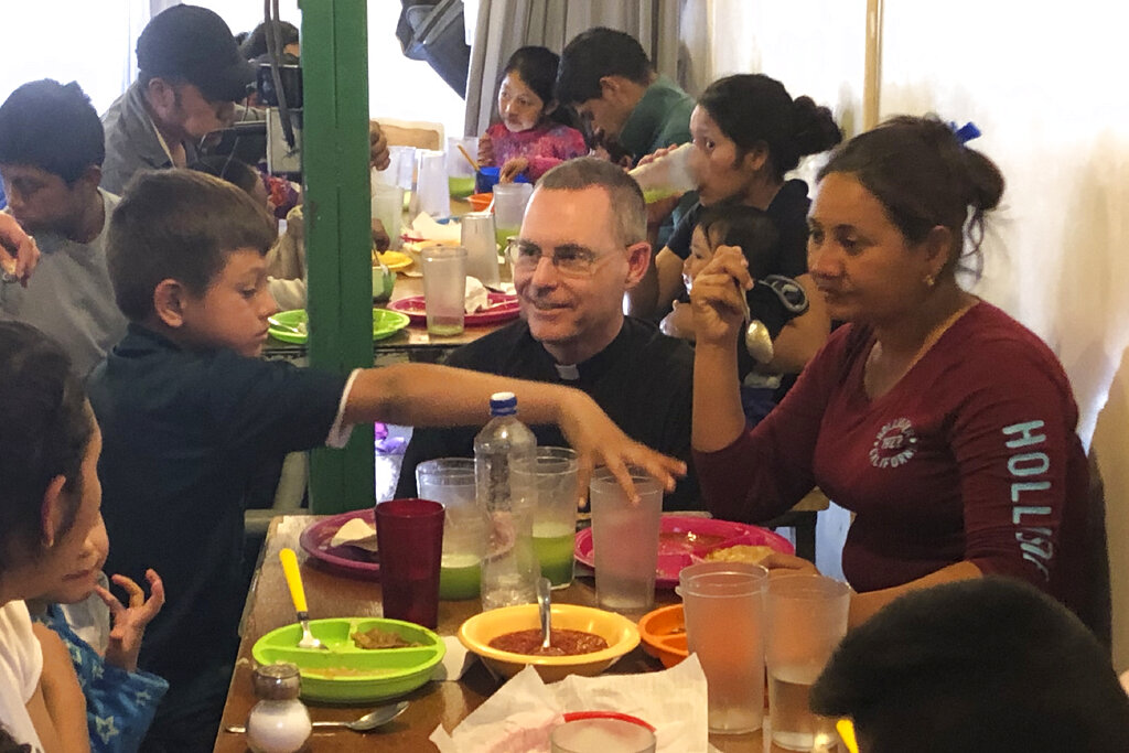 In this Feb. 20, 2020 photo, Jesuit priest Sean Carroll joins a group of asylum-seekers from Honduras in the cafeteria of a migrant-outreach center that his organization, the Kino Border Initiative, operates near the U.S.-Mexico border in Nogales, Mexico. Before the coronavirus gained global attention, Carroll's agency opened the facility just inside the Mexican border. Carroll — who works full-time in Mexico — hoped to expand a twice-daily meal service but now amid worries about COVID-19, neither venue is being used as a dining hall. Instead, migrants line up outside the two buildings and approach the doors one at a time. (AP Photo/David Crary)
