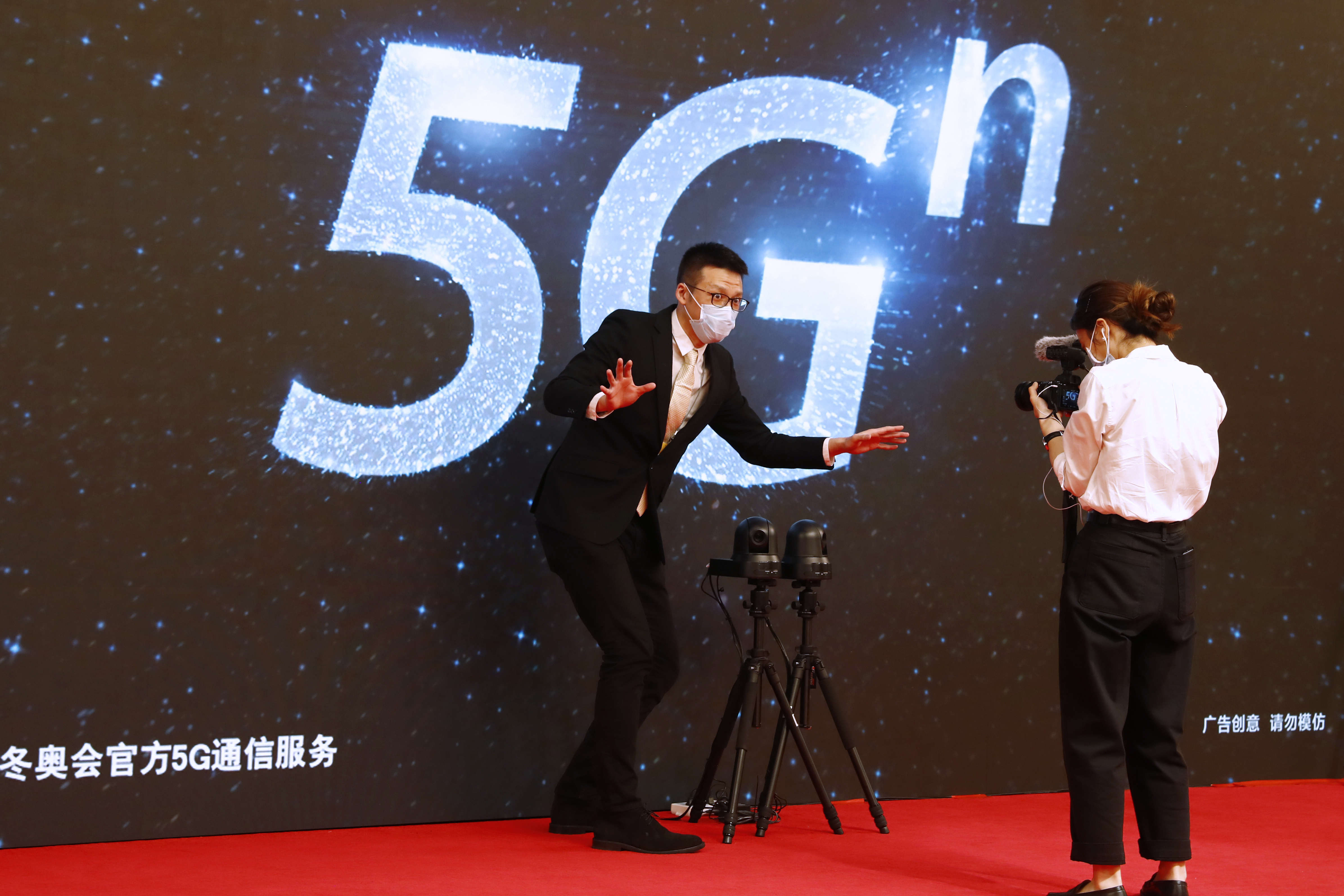 A local media reporter wearing a face mask following the coronavirus outbreak explains for the camera the technology involved in broadcasting the news conference by Guo Weimin, spokesman for the National Committee of the Chinese People's Political Consultative Conference (CPPCC), in front of a 5G sign at a media center in Beijing, China Wednesday, May 20, 2020. (Thomas Peter/Pool Photo via AP)