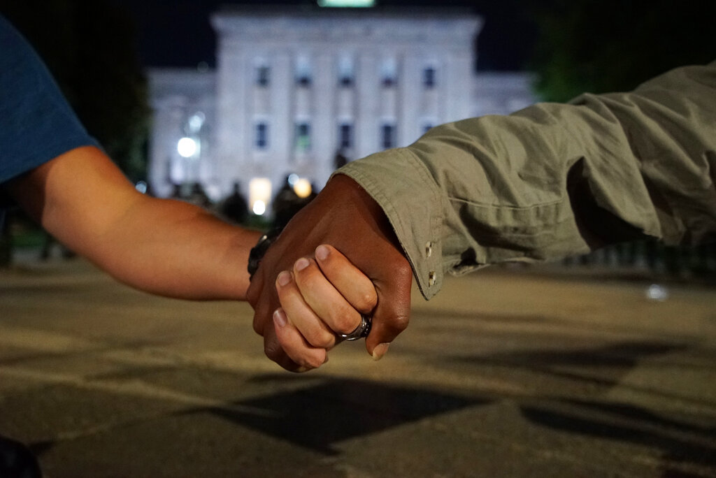 A white man and a black man clasp hands as police in the background guard the old state capitol in Raleigh, N.C., on Monday, June 1, 2020. It was the second day of protests in the North Carolina capital following the death of Minnesotan George Floyd while in police custody. (AP Photo/Allen G. Breed)