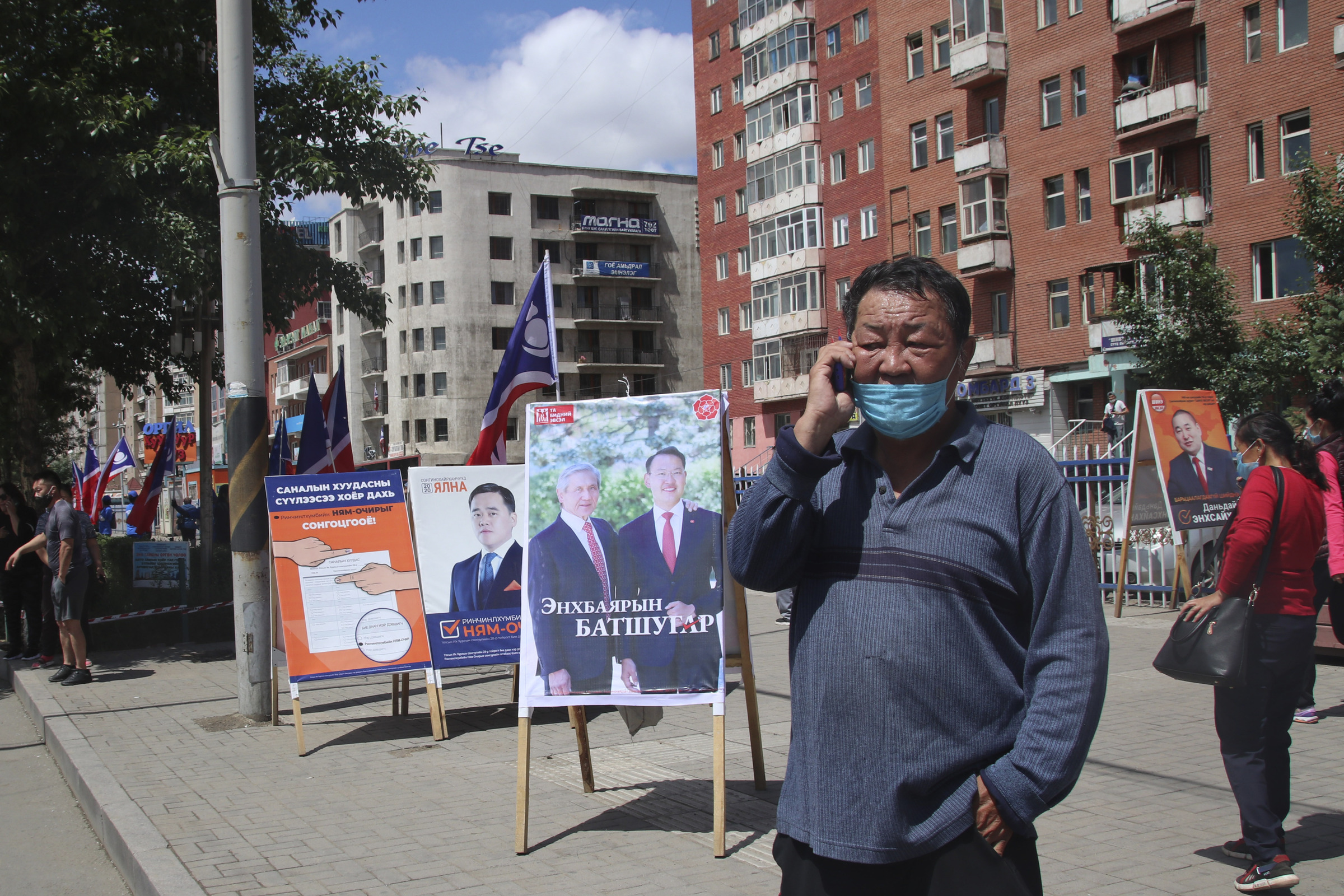 A man wearing a mask and using his phone stands near the election stand of Mongolian People's Revolutionary Party or MPRP featuring ex-president of Mongolia Enkhbayar Nambar and his son Batshugar in the Songinokhairkhan district on the outskirt of Ulaanbaatar, Mongolia, Monday, June 22, 2020. Mongolia holds parliamentary elections on Wednesday, continuing a nearly 30-year democratic system in a vast but lightly populated country sandwiched between authoritarian regimes in Russia and China and beset by economic problems. (AP Photo/Ganbat Namjilsangarav)