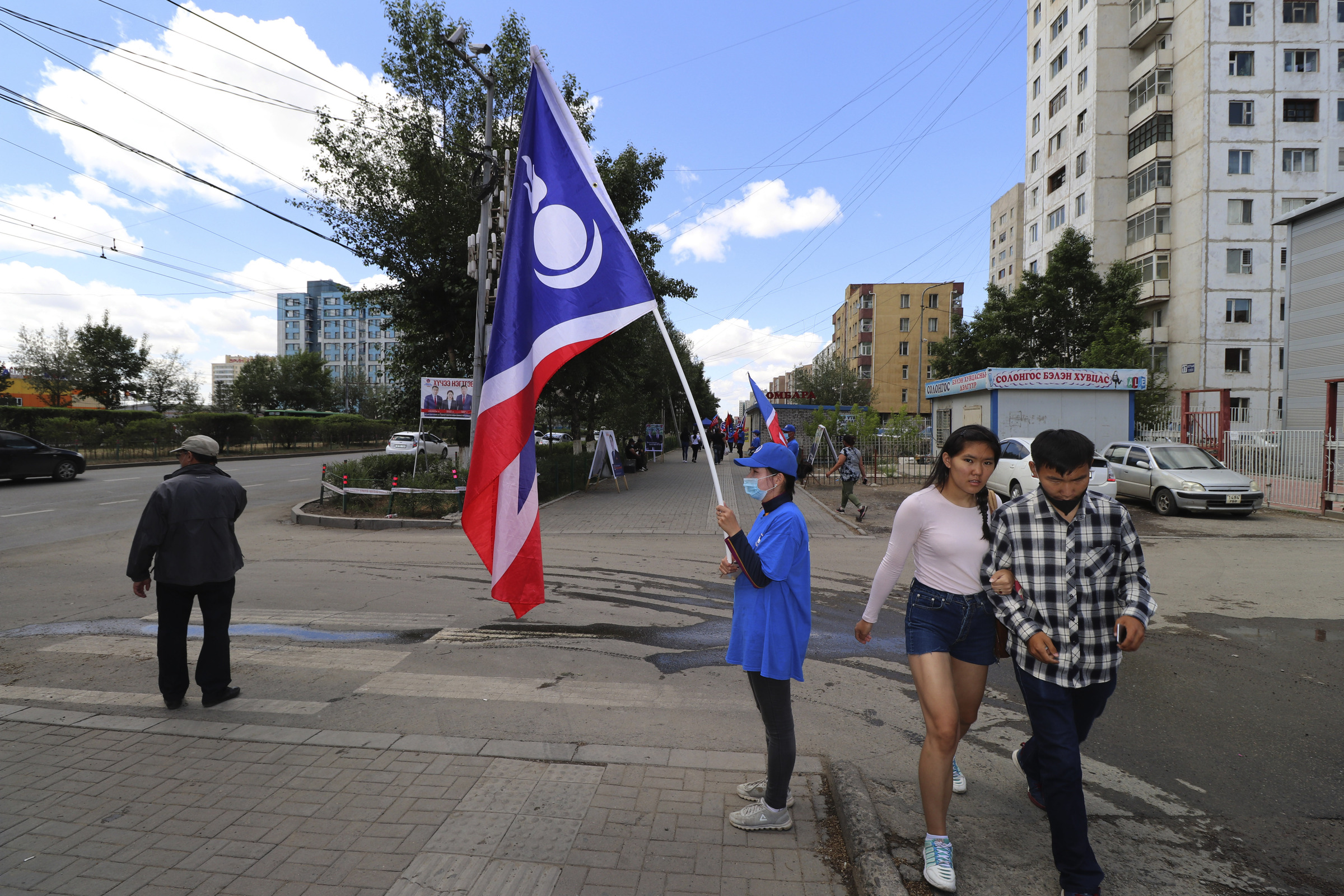 A couple walks past a Democratic Party campaign worker holding the party flag in the Songinokhairkhan district on the outskirts of Ulaanbaatar, Mongolia, Monday, June 22, 2020. Mongolia holds parliamentary elections on Wednesday, continuing a nearly 30-year democratic system in a vast but lightly populated country sandwiched between authoritarian regimes in Russia and China and beset by economic problems. (AP Photo/Ganbat Namjilsangarav)