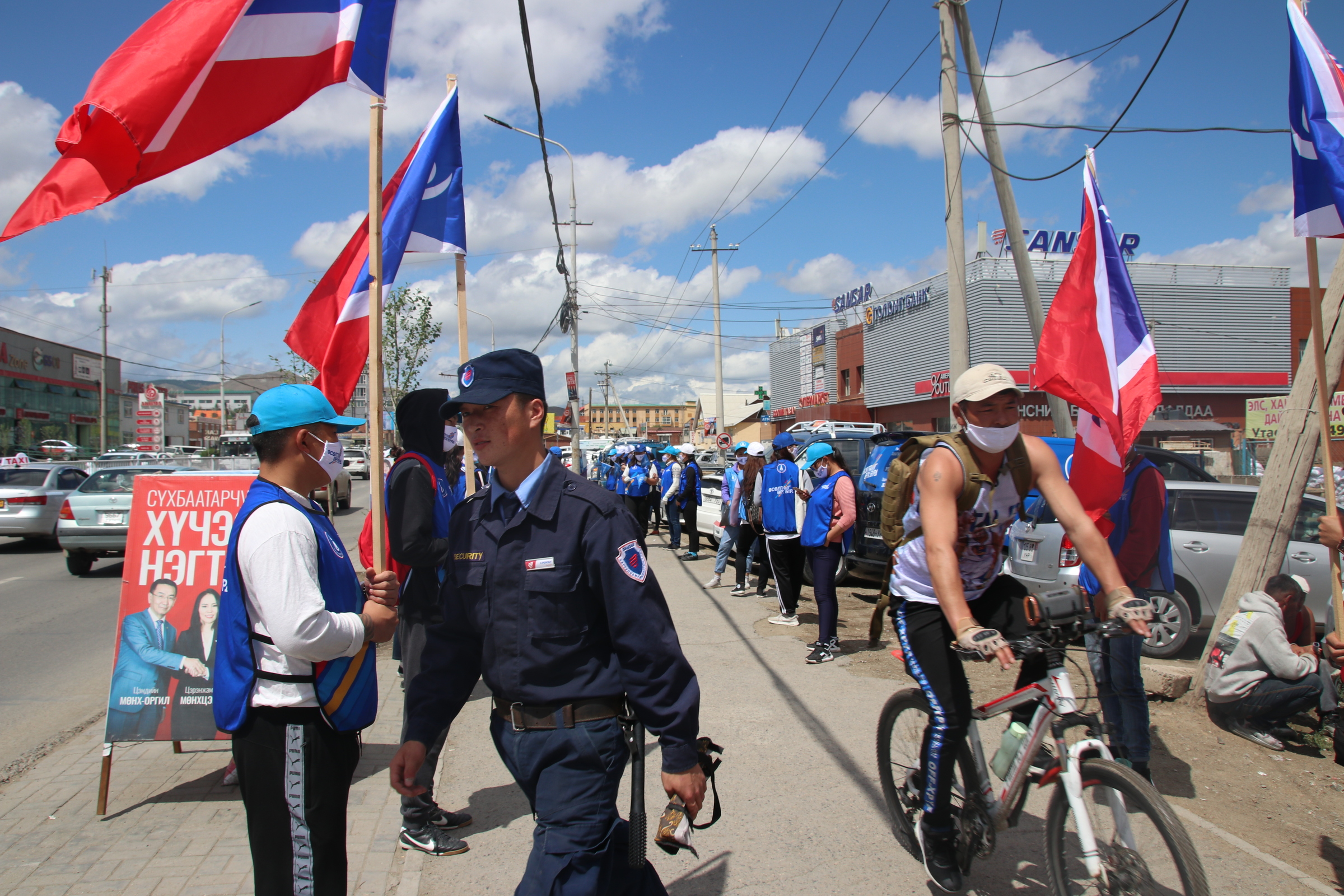 A security guard and a cyclist past by election workers of the opposition Democratic Party campaigning in the Chingeltei district, northern outskirts of Ulaanbaatar, Mongolia, Monday, June 22, 2020. Mongolia holds parliamentary elections on Wednesday, continuing a nearly 30-year democratic system in a vast but lightly populated country sandwiched between authoritarian regimes in Russia and China and beset by economic problems. (AP Photo/Ganbat Namjilsangarav)