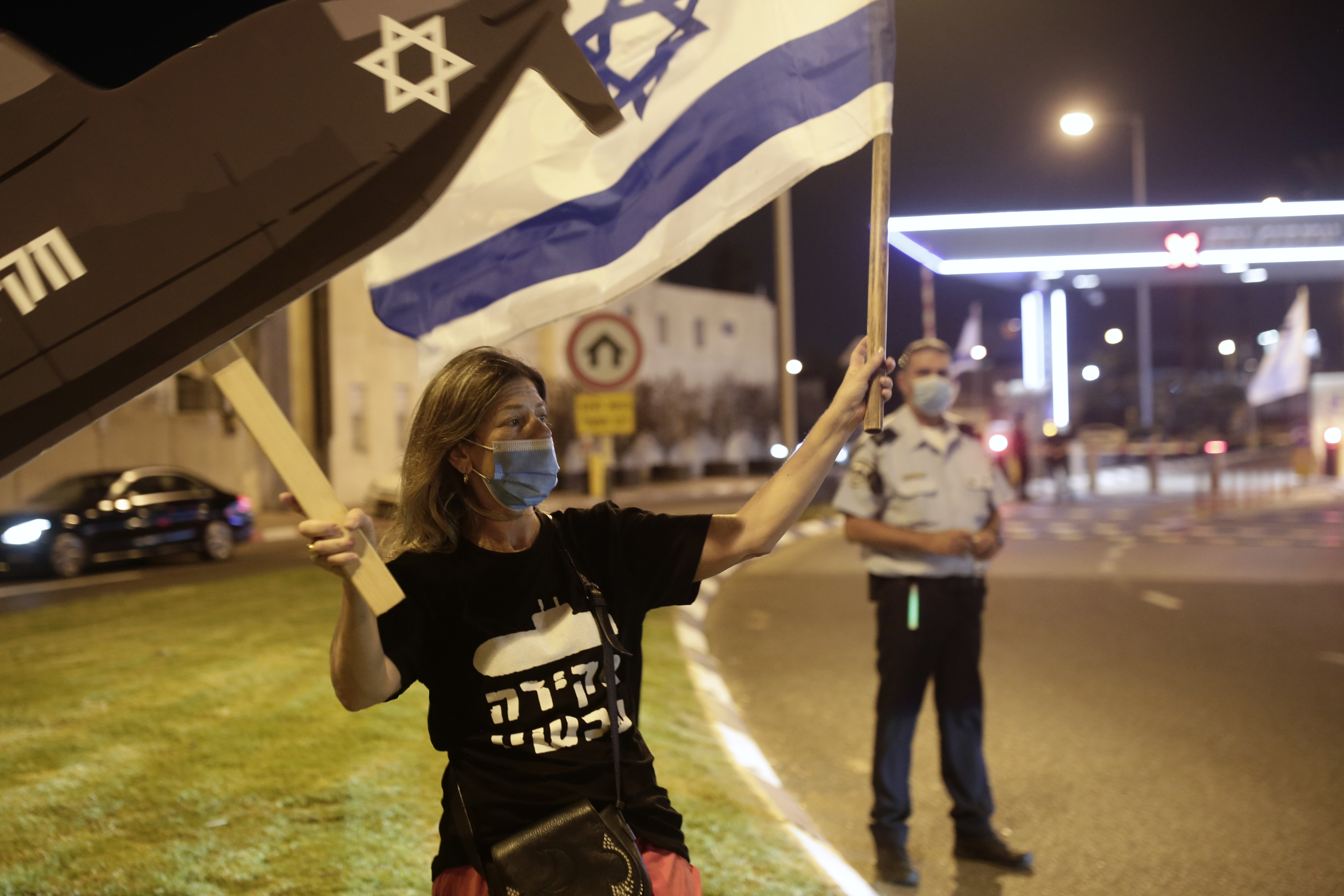 A protester waves an Israeli flag and a placard as a policeman watches a demonstration to block an entrance to Ben Gurion Airport, where Prime Minister Benjamin Netanyahu and his family were expected to fly with an Israeli delegation to the U.S. for a ceremony with the United Arab Emirates, in Tel Aviv, Sunday, Sept. 13, 2020. (AP Photo/Maya Alleruzzo)