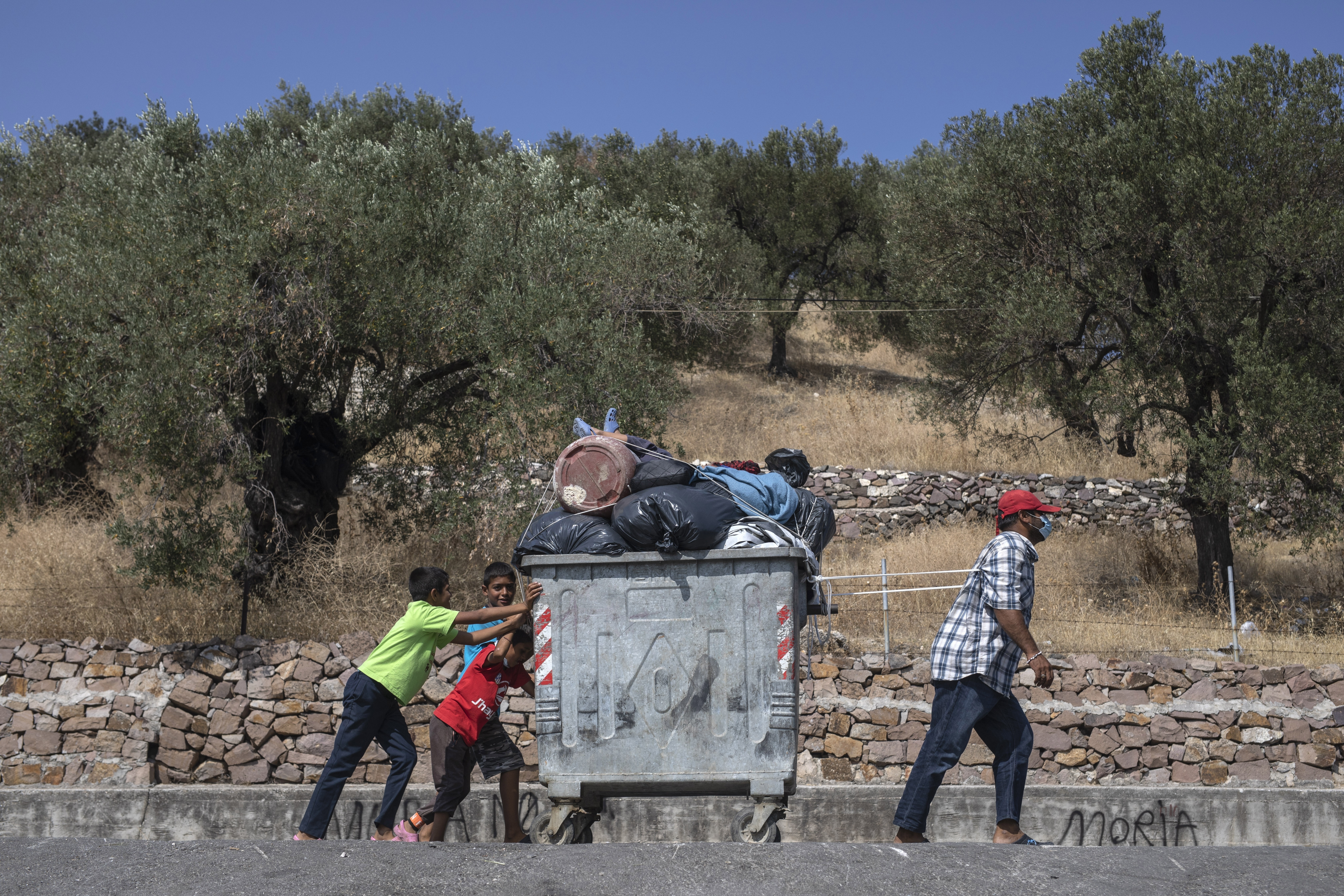 Migrants use a trash bin to move their belongings from the burned Moria refugee camp to a new army-built facility in Kara Tepe on the northeastern island of Lesbos, Greece, Friday, Sept. 18, 2020. Police on the Greek island of Lesbos on Friday resumed relocating migrants rendered homeless when fires ravaged the country's largest refugee camp amid a local COVID-19 outbreak. (Foto AP Photo/Petros Giannakouris)