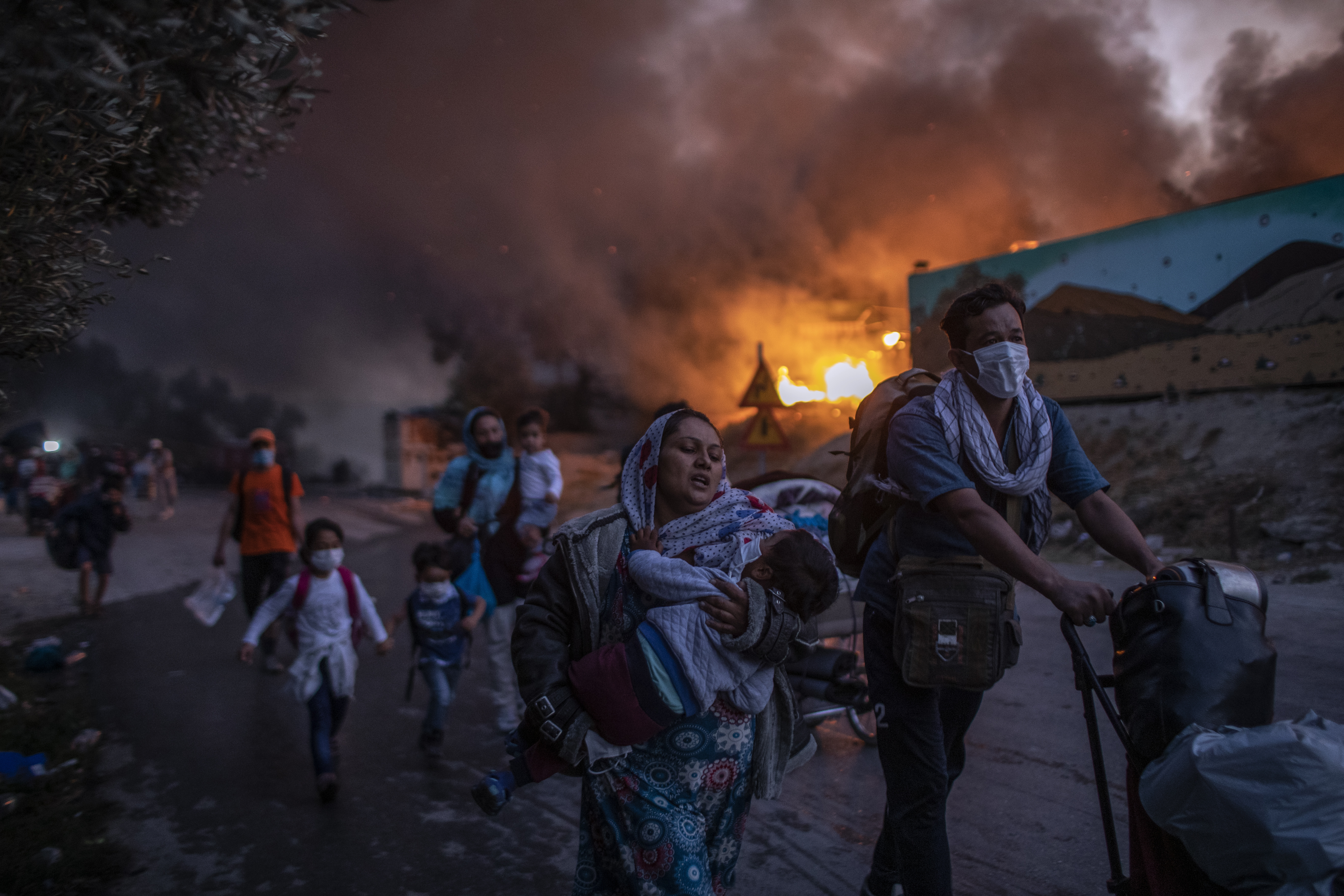 Refugees and migrants carrying their belongings flee a fire burning at the Moria camp on Lesbos island, Greece, Wednesday, Sept. 9, 2020. Moria refugee camp was never far from crisis. Created in the wake of a massive influx of migrants five years ago, Greece's largest refugee facility quickly exceeded capacity, spilling into surrounding olive groves on the island of Lesbos. The camp's life ended as it began, in drama: Successive fires that started before dawn on Sept. 9 devastating the site and making 12,000 inhabitants homeless during a COVD-19 lockdown. (AP Photo/Petros Giannakouris)