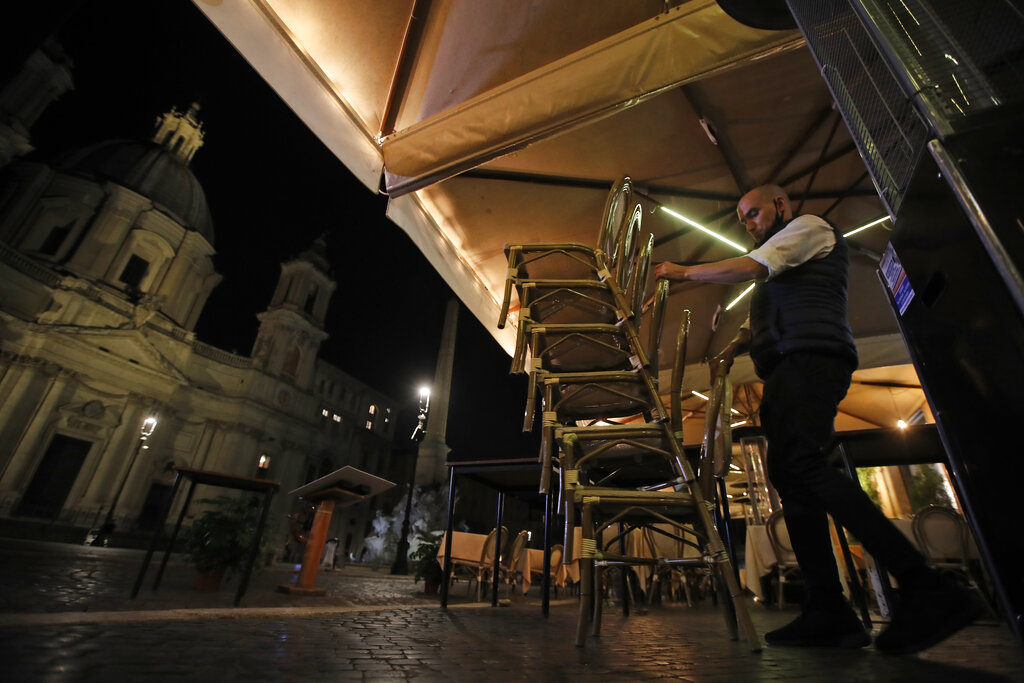 A waiter collects chairs at a cafe restaurant in Piazza Navona Square before the start of a curfew, Friday, Oct. 23, 2020. Italian Premier Giuseppe Conte, who imposed severe-stay-at-home limits on citizens early on, then gradually eased travel and other restrictions, has been leaving it up to regional governors in this current surge of infections to order restrictions such as overnight curfews, including in places like Rome, Milan and Naples. (AP Photo/Alessandra Tarantino)