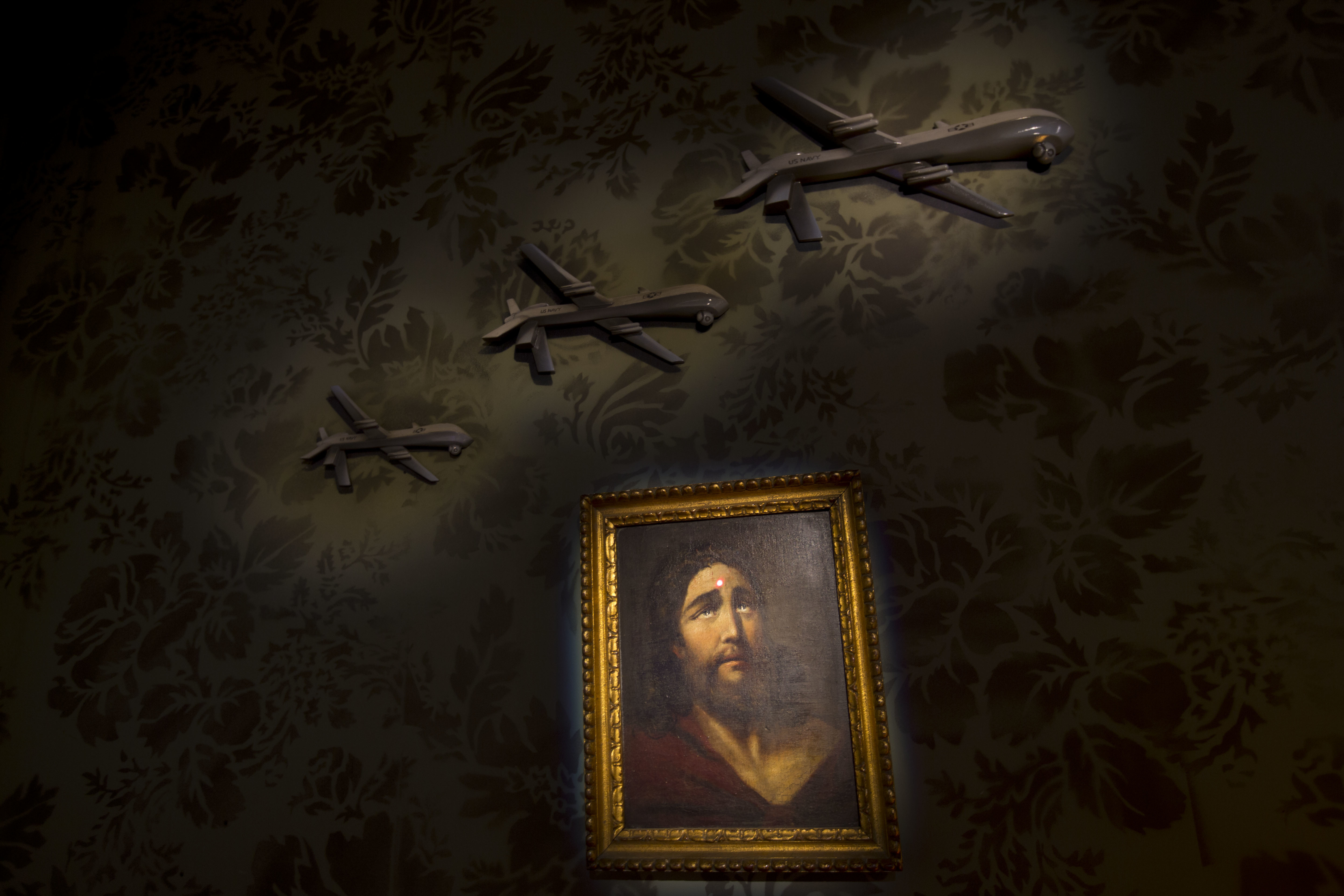 A wall decorated with models of drones and a painting of Jesus with a sniper's dot on his forehead is seen in the 